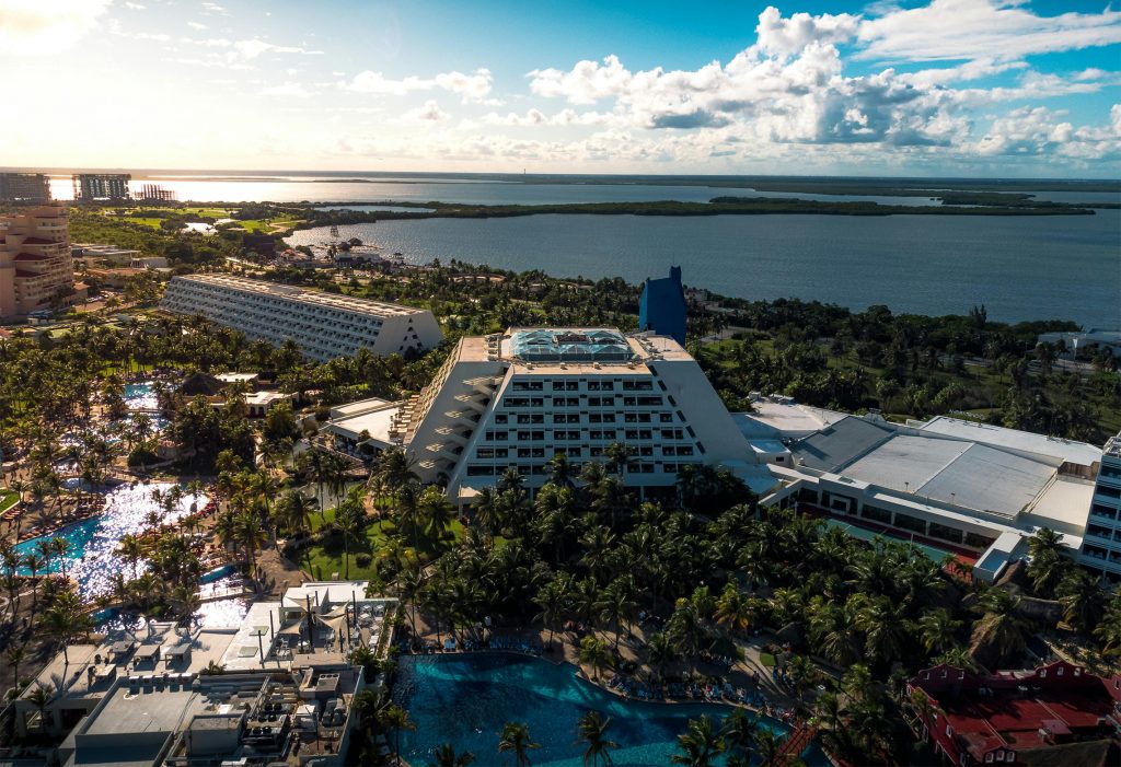 Aerial view of a coastal Cancun resort featuring a large, pyramid-shaped hotel surrounded by lush greenery and several swimming pools. The ocean stretches in the background under a partly cloudy sky.