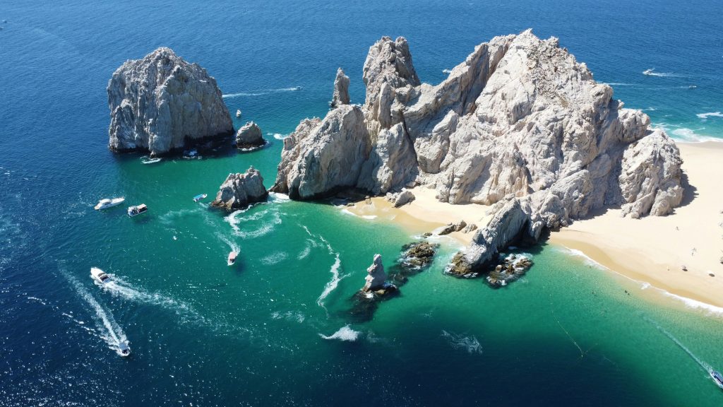 Aerial view of dramatic rock formations on a sandy beach surrounded by turquoise and deep blue water, with several boats leaving white trails near the shore—an unforgettable Cabo High School Trip destination.