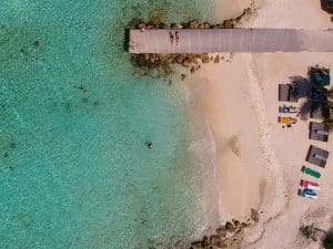 Aerial view of a tranquil beach scene with clear turquoise water. A wooden pier extends over the water, where two people relax. Sunbeds and umbrellas are scattered on the sandy shore, and a few swimmers enjoy the pristine sea.