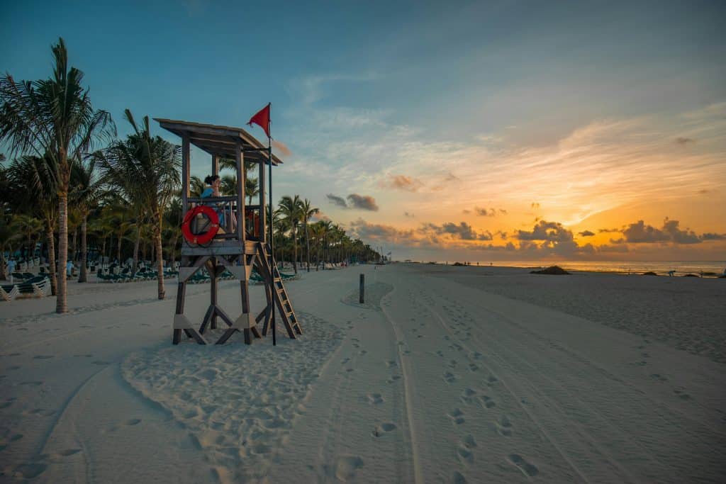 Lifeguard tower on a sandy beach during sunrise. A red flag and lifebuoy are attached to the tower. Palm trees line the beach, and sun loungers are visible in the background. The sky displays warm colors and scattered clouds.