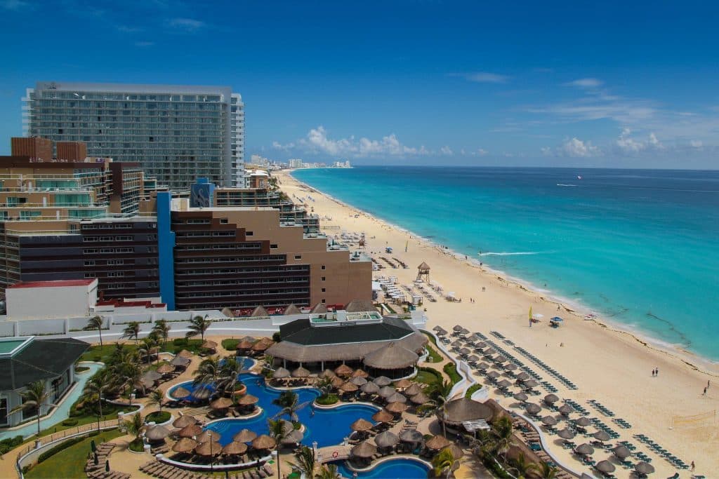 Aerial view of a beachfront resort in Cancun with high-rise hotels, multiple pools, palm trees, and sun loungers. The turquoise sea and sandy beach stretch into the distance under a clear blue sky.