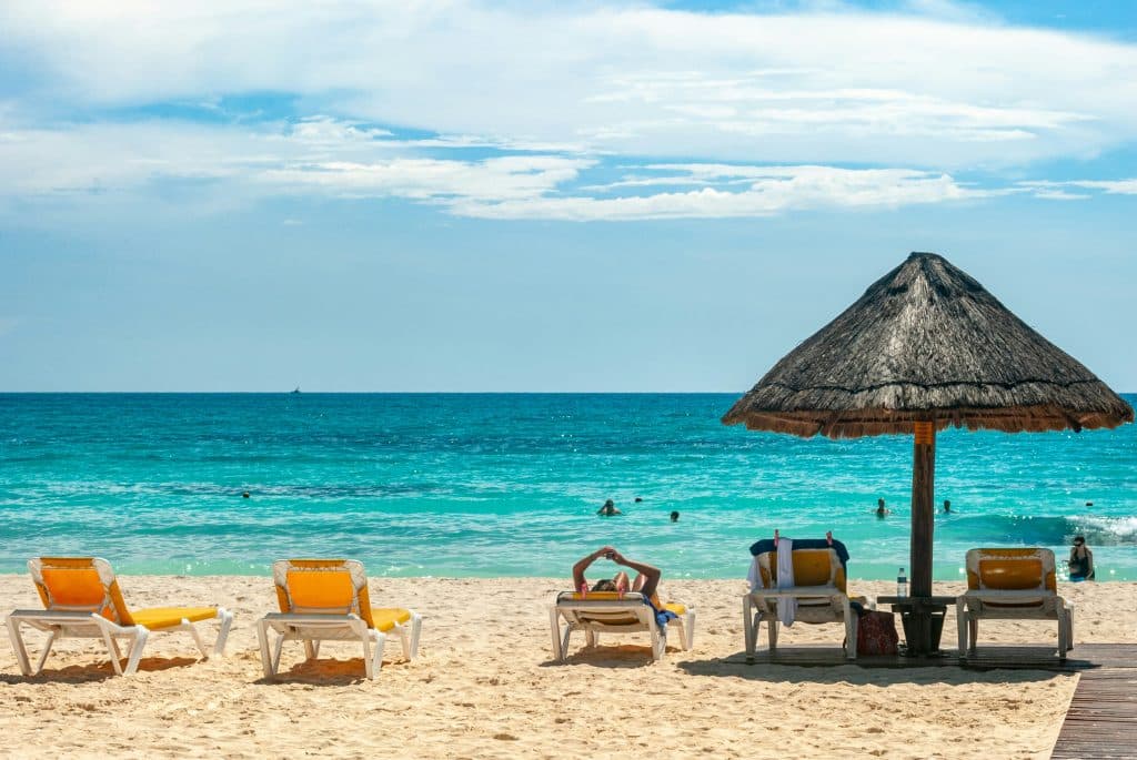 Five lounge chairs and a thatched umbrella sit on a sandy beach facing turquoise ocean waves. People swim in the water under a partly cloudy blue sky. The scene looks relaxing and tropical.