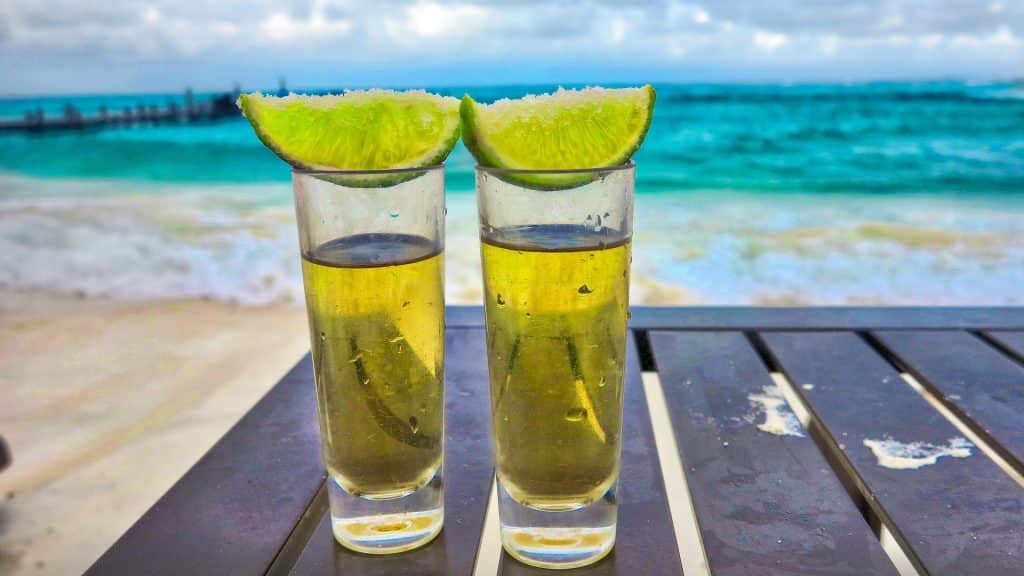 Two shot glasses filled with golden liquid and topped with lime wedges sit on a table, with a sandy beach and turquoise ocean waves in the background.