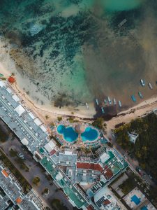 Aerial view of a resort with two large blue pools near a sandy beach and clear, shallow ocean water. Several small boats are lined up along the shore, and buildings surround the pool area.
